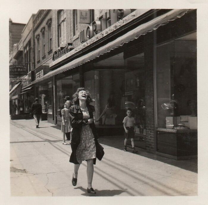 Black and white historical photo of a joyful woman walking on a city sidewalk with children in the background.