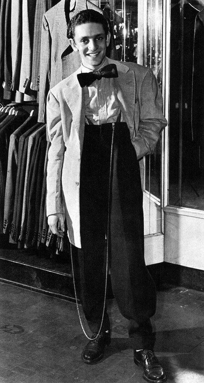 Young man wearing exaggeratedly oversized pants and a bow tie, standing in a vintage clothing store, historical photo.