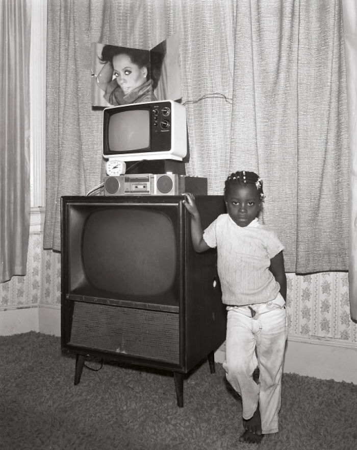 Young girl standing next to vintage television sets in a living room, a classic scene from historical photos.