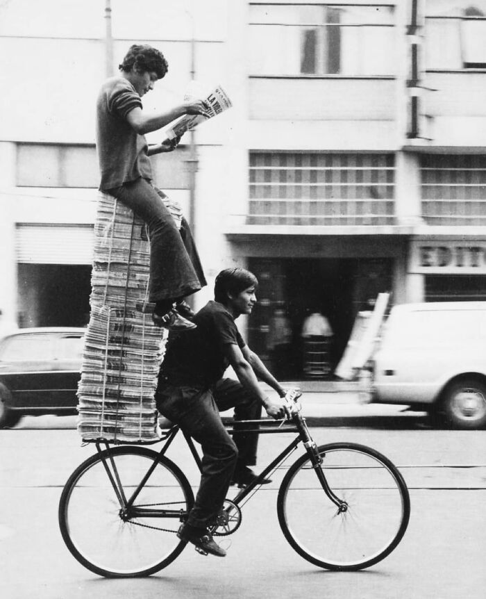 Black and white historical photo of two men transporting newspapers on a bicycle in a busy city street.