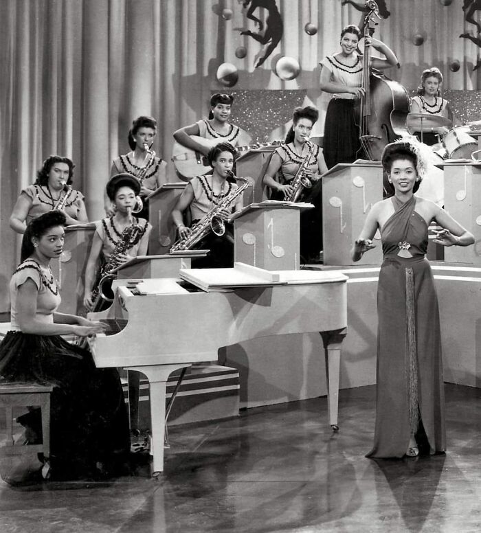 Black and white historical photo of an all-female jazz band performing with piano, saxophones, guitar, and vocalist on stage.