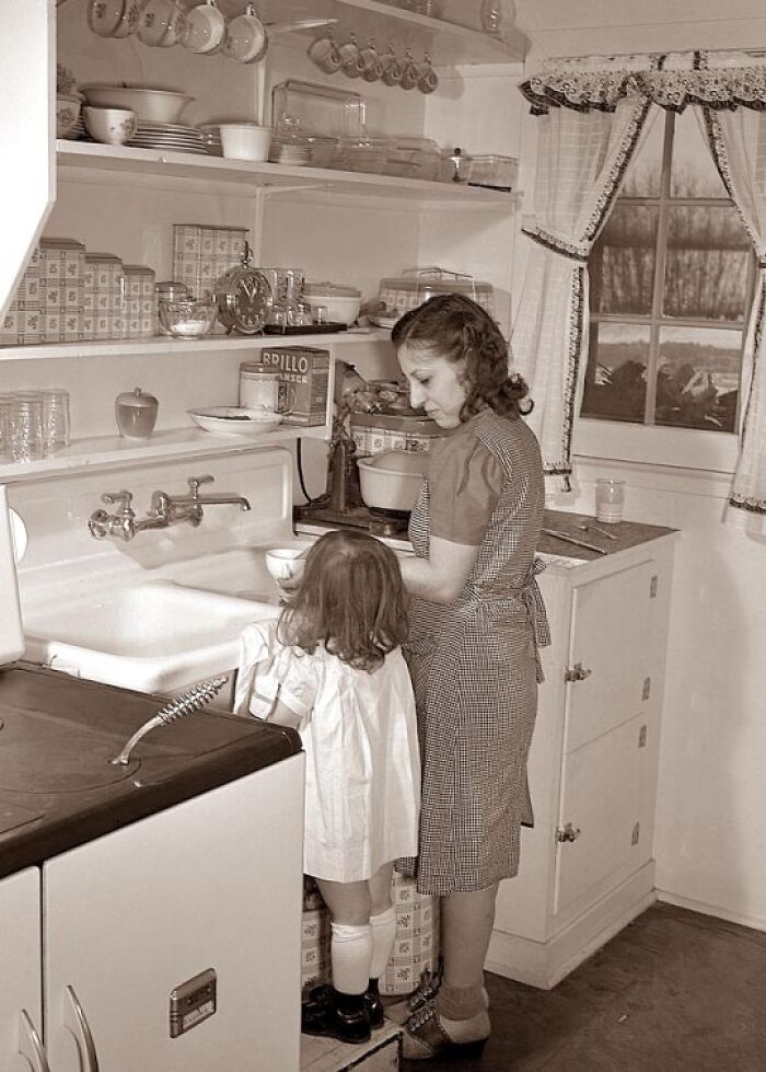 Vintage historical photo of a woman and child in a 1940s kitchen preparing food together by the sink.