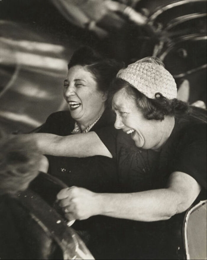 Two women laughing joyfully while riding an amusement park car in a historical photo from the past.