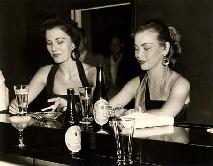 Two women in vintage dresses sitting at a bar with beer bottles and glasses in a historical photo.