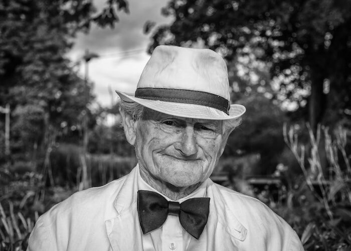 Older man wearing a white hat and black bow tie smiling outdoors, representing the coolest person to have ever lived.
