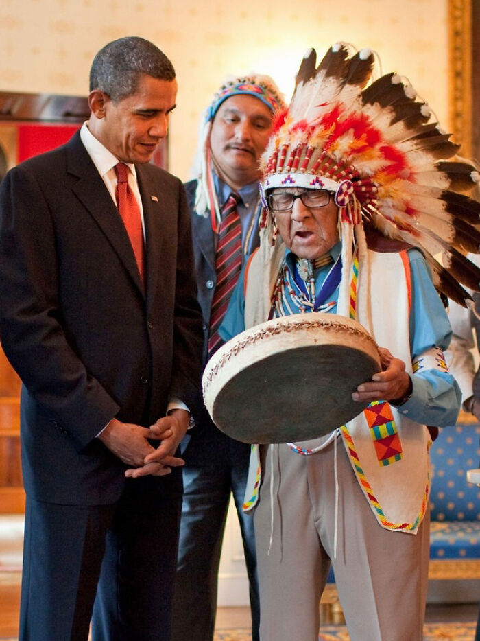 Barack Obama listens respectfully to a Native American elder playing a drum, highlighting the coolest person to have ever lived.