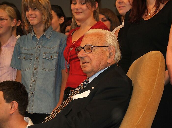 Elderly man in glasses and suit sitting in chair surrounded by smiling young people sharing coolest person moments online