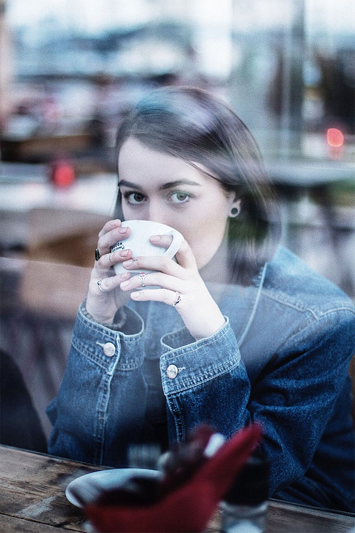 Young woman in denim jacket drinking coffee inside a café, reflecting on what not to do when visiting the US.