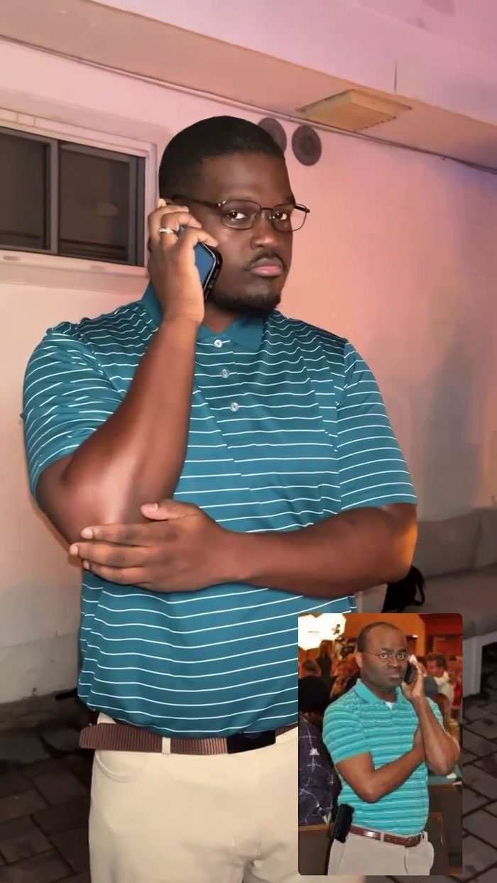 Man at meme-themed party holding phone, wearing striped shirt and glasses; understood the assignment perfectly.