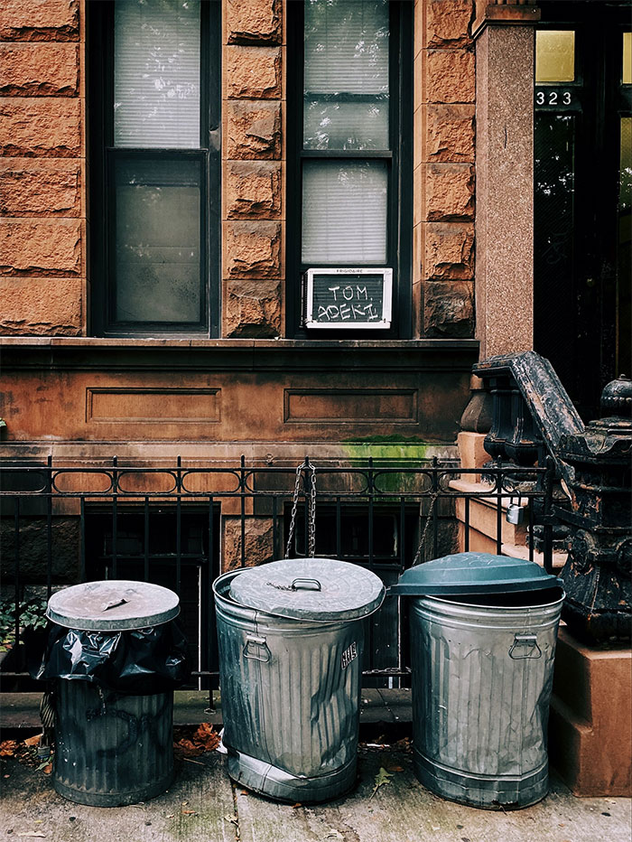 Three metal trash cans with lids outside a brownstone building in a US urban neighborhood, highlighting what not to do.