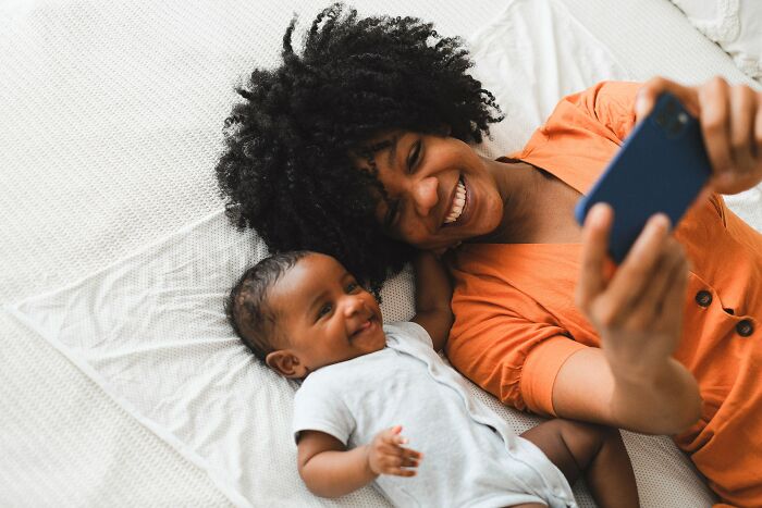 A smiling mother and baby lying on a bed taking a selfie, illustrating unspoken mom code moments.