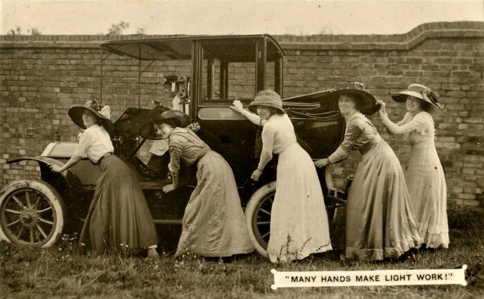 Five women in vintage dresses and hats push an old car, illustrating teamwork and the coolest person concept.