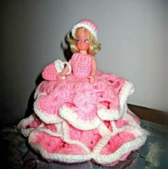 A doll in a pink and white crochet dress sits on a dark surface, humorously highlighting the theme of getting old.
