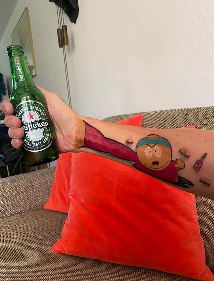 A Heineken bottle held by a tattooed arm featuring a cartoon design, with a couch and orange cushions in the background.
