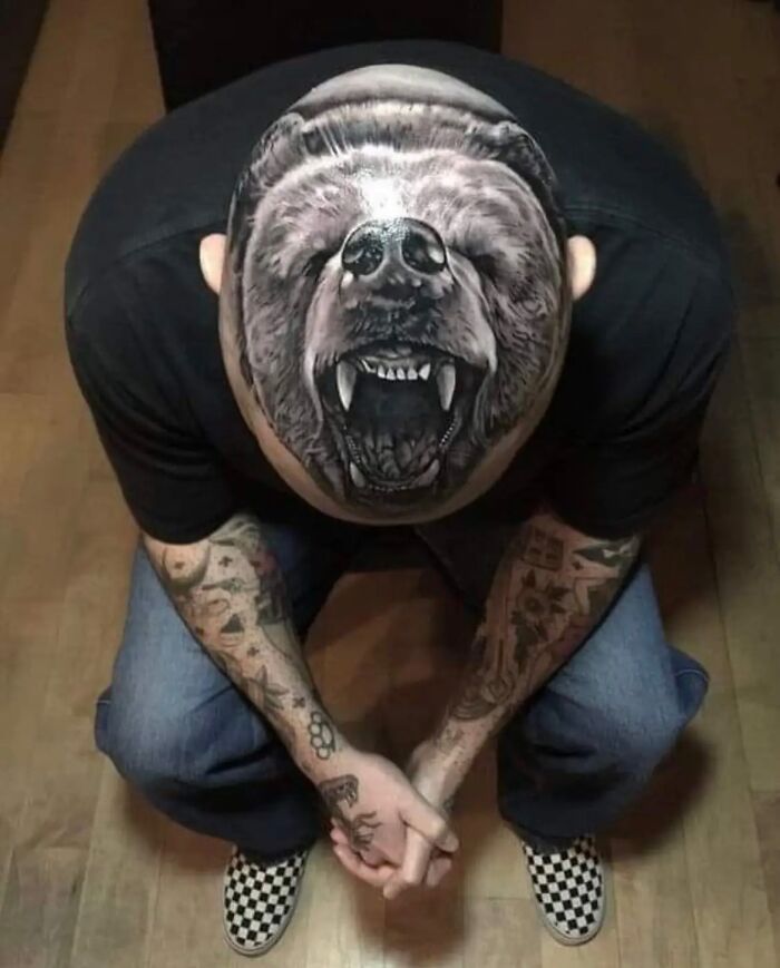 Person with tattoos, including a large bear tattoo on their head, sitting with hands clasped, wearing checkered shoes.