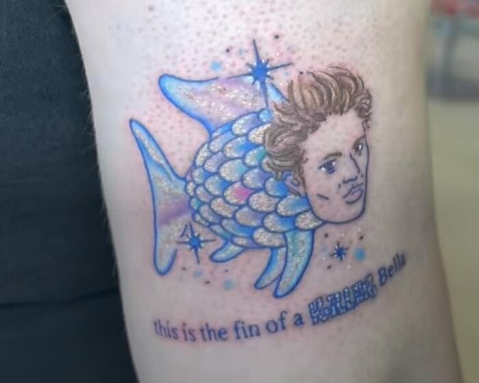 Tattoo of fish with a human face and glittery scales on skin, featuring humorous text below the design.