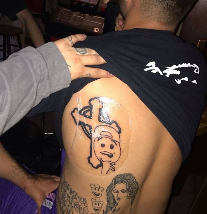 Unusual tattoo design on a man’s back, featuring a thinking emoji with crossbones.