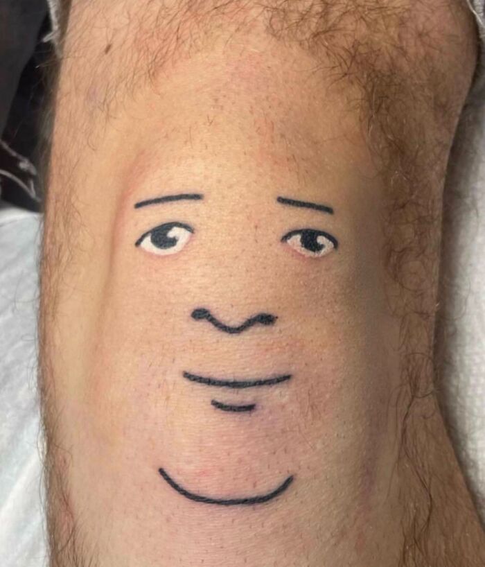 Minimalist face tattoo on a knee with simple lines for eyes, mouth, and nose.
