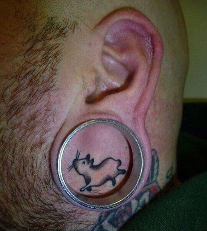 Unusual ear tattoo featuring a small black rat design within a large gauge earlobe piercing.