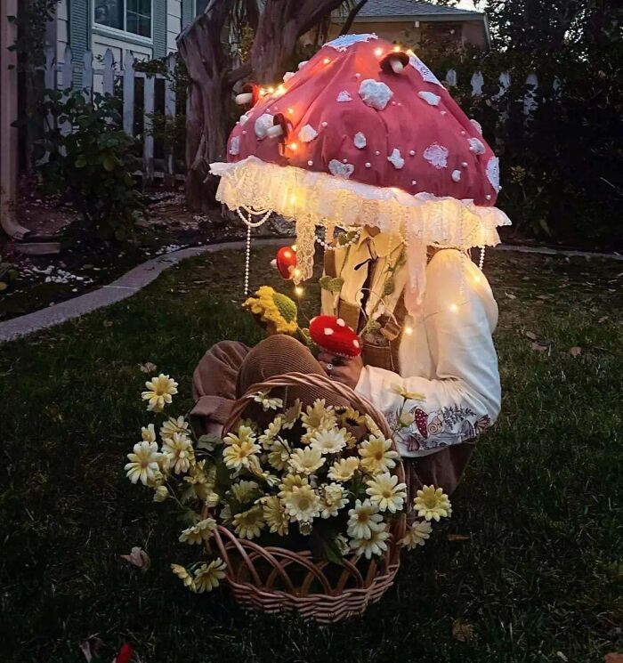 Person wearing a clever Halloween costume resembling a glowing red mushroom with white spots sitting beside a basket of flowers.