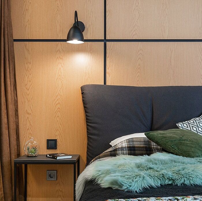Modern bedroom wall decor with wood paneling, dark cushioned bed, soft blankets, and a bedside table under a black wall lamp.
