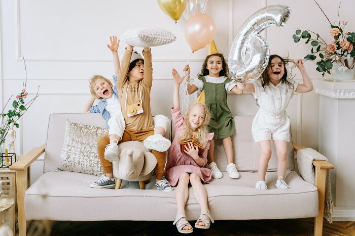 Five children celebrating a birthday party on a couch with balloons and a large number six balloon in a living room.