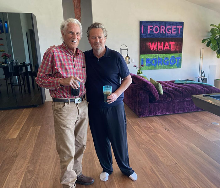 Two men standing in a living room, holding drinks, with colorful artwork in the background.