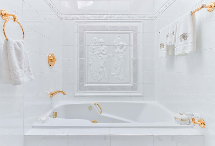 All-white ceramic bathroom featuring elegant wall tile design and golden accessories around the bathtub area.