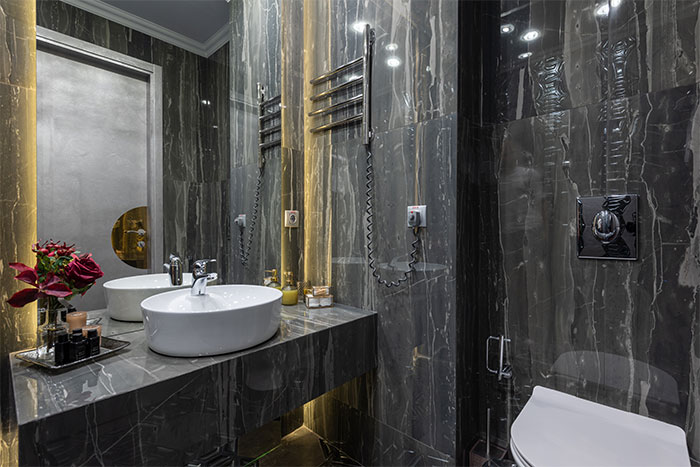 Modern bathroom featuring large black bathroom tiles on walls and flooring with a gray stone sink and illuminated mirror.