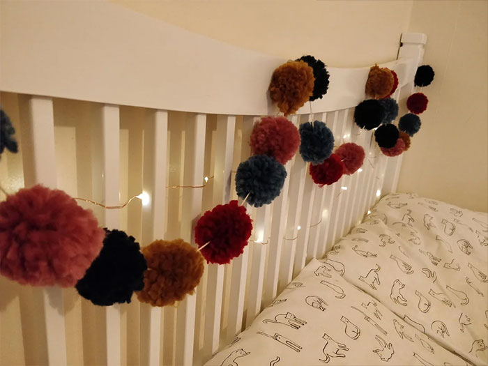 Colorful pom pom garland hanging on white bed frame as a creative bedroom wall decor idea with soft lighting.