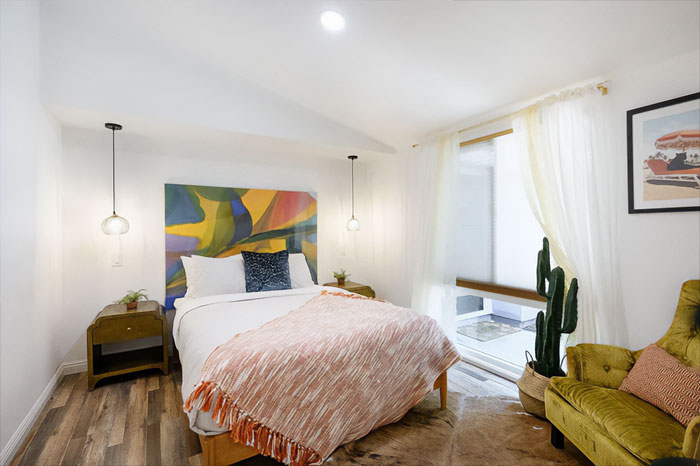 Modern bedroom interior featuring colorful wall decor, cozy bedding, pendant lights, and natural light through sheer curtains.