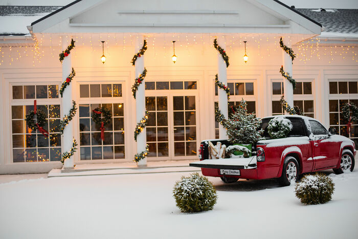 Red car with Christmas tree and affordable Christmas decor outside a house decorated with lights and garlands in the snow.