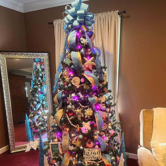 Decorated Christmas tree with blue bow topper, seashell and starfish ornaments, and glowing purple lights for festive decor.