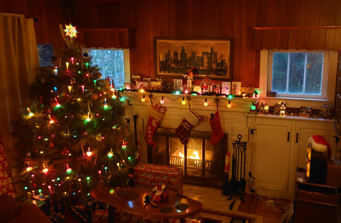 Decorated Christmas tree with colorful lights and ornaments next to a cozy fireplace with hanging stockings.