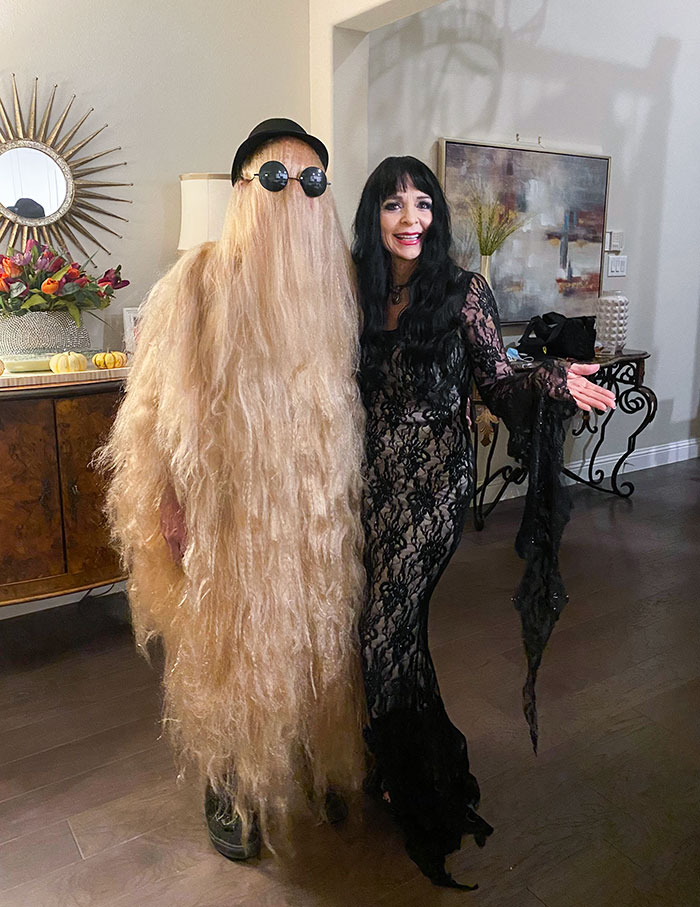 Two people in clever Halloween costumes, one with a long hair disguise and the other in a black lace dress indoors.