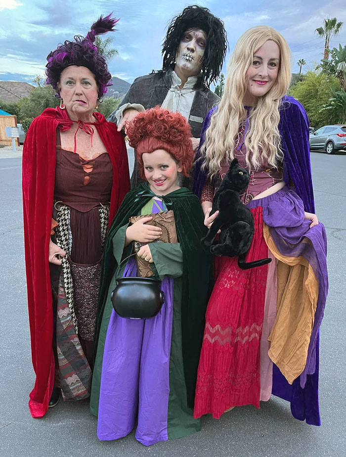 Group of four people in clever Halloween costumes inspired by witches and spooky characters holding a black cat outside.