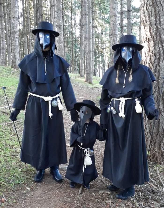 Family dressed in clever Halloween costumes as plague doctors with black robes, beak masks, and hats in a forest setting.