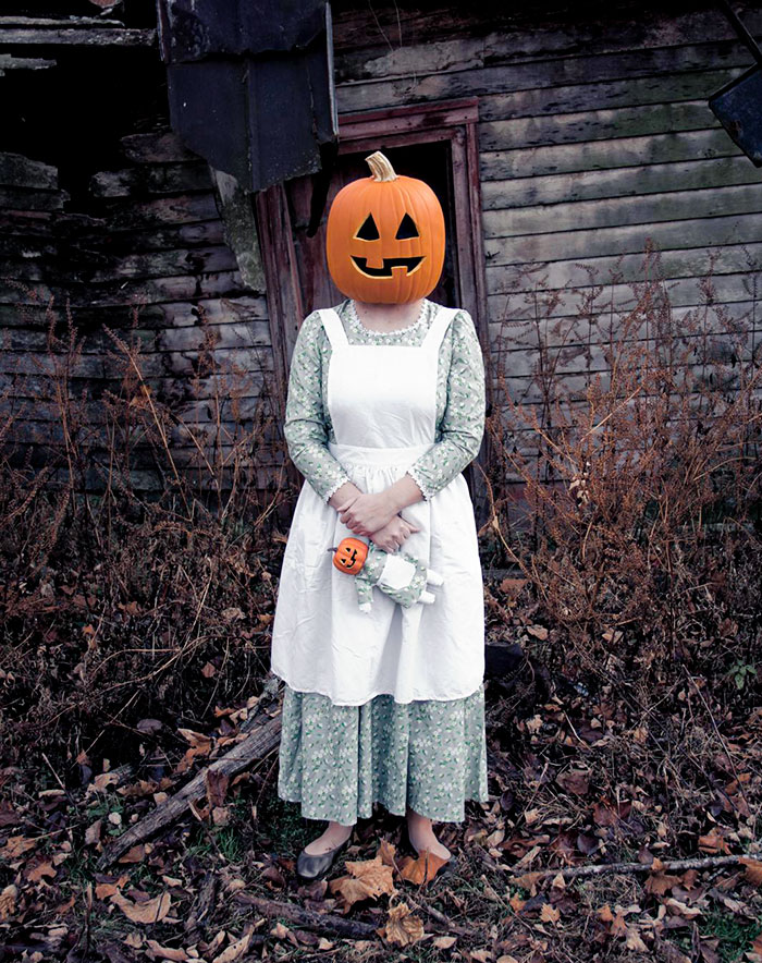 Person wearing clever Halloween costume with a pumpkin head and vintage dress standing outdoors near old wooden building.