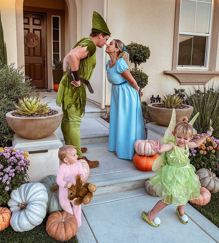 Family dressed in clever Halloween costumes including Peter Pan, Wendy, and fairy with pumpkins on porch steps.