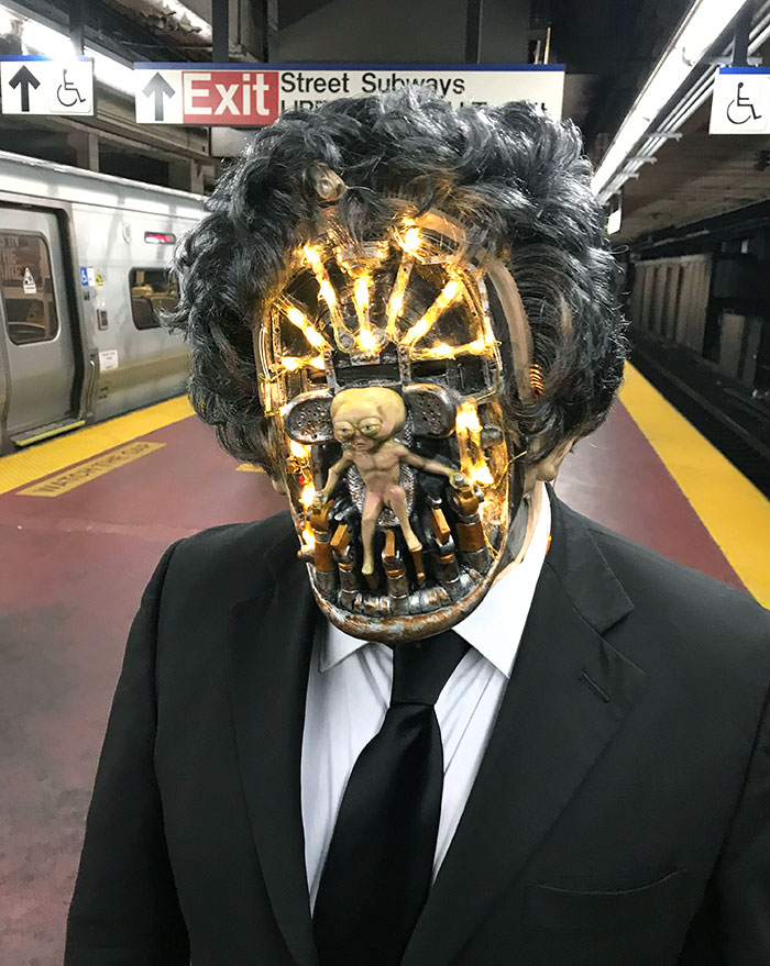 Man in a clever Halloween costume with illuminated mask and small alien figure on subway platform, wearing black suit and tie.