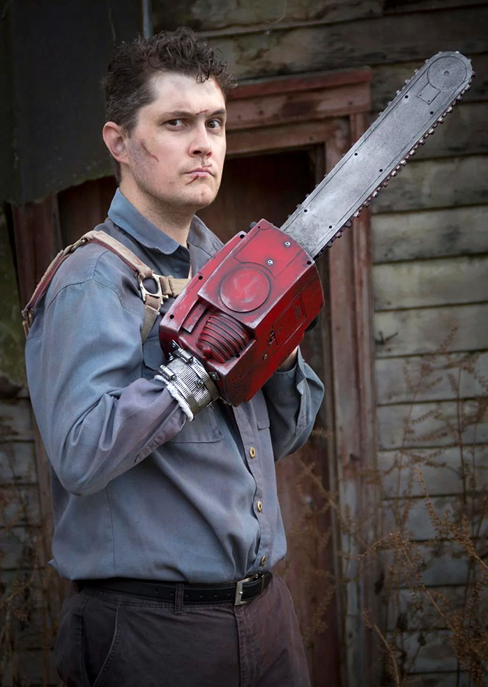 Man in clever Halloween costume holding a large prop chainsaw, standing in front of an old wooden building.