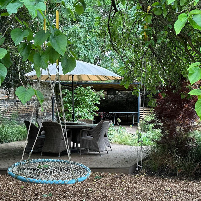 Backyard deck with wicker chairs, round table under a large umbrella, surrounded by lush trees and garden greenery.