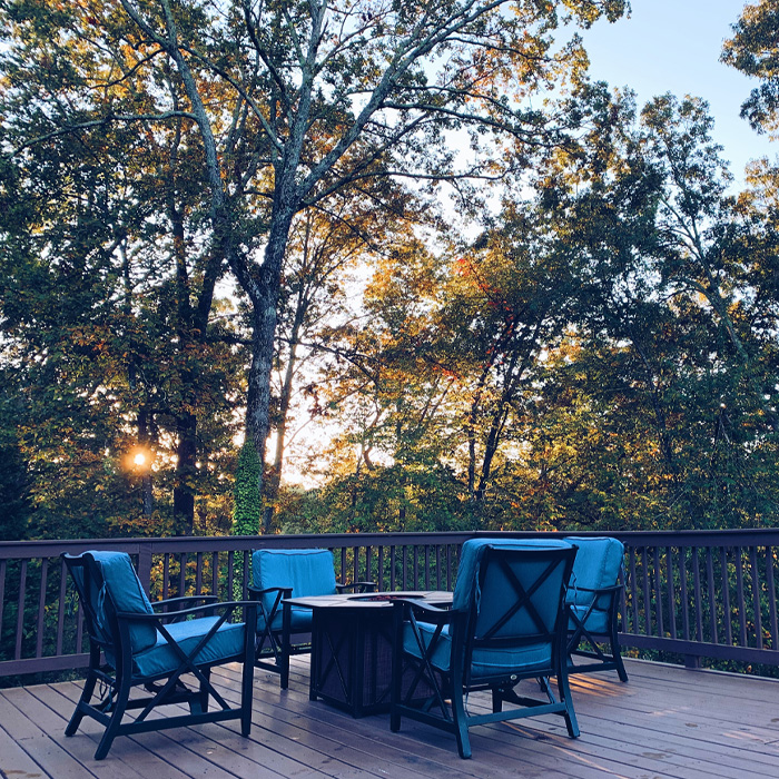 Fire pit on a backyard deck surrounded by blue cushioned chairs during sunrise with trees in the background.