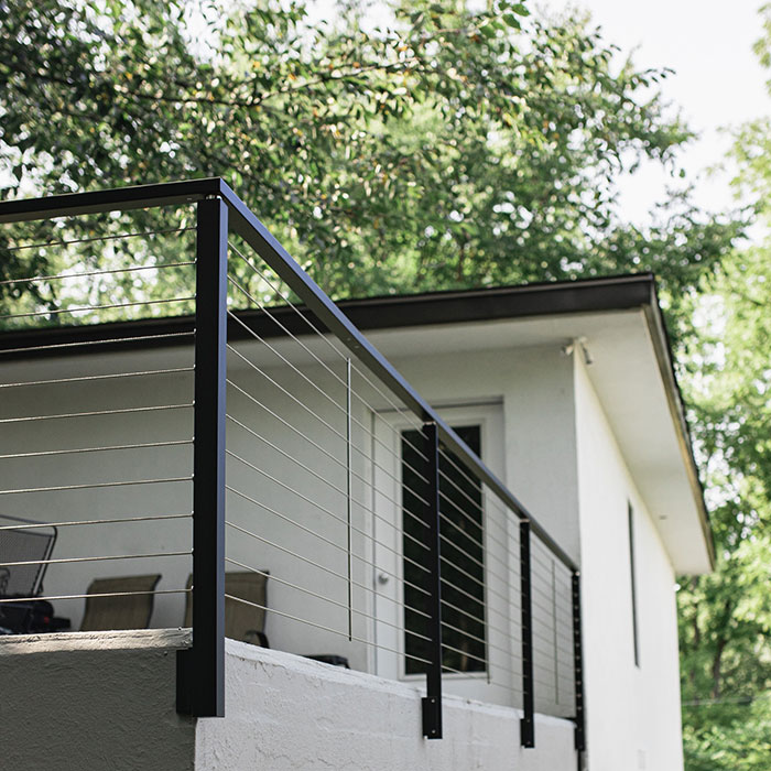 Black deck railing with horizontal cables on a white house exterior surrounded by green trees in daylight