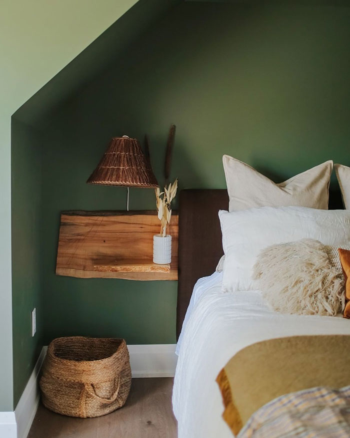 Raw wooden floating nightstand mounted on green bedroom wall next to bed with white pillows and woven basket below.