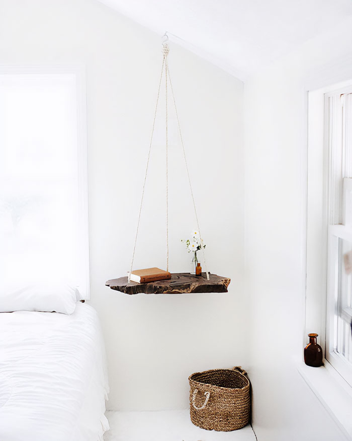 Hanging modern floating nightstand made from natural wood slab with books and flowers in a bright minimalist bedroom.