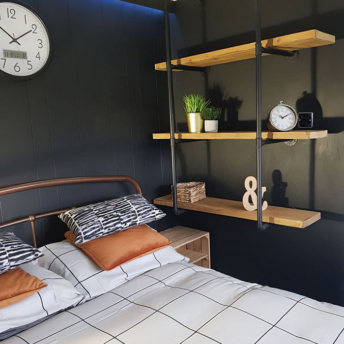 Floating pipe nightstand with wooden shelves in a modern bedroom featuring black walls and minimalist decor.