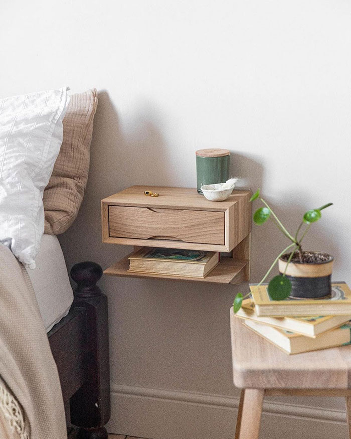 Double decker modern floating nightstand in natural wood mounted beside bed with small decor and books below shelf.