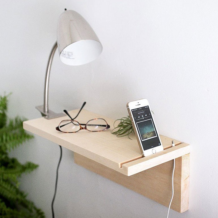 Floating nightstand with phone slot holding a charging smartphone, glasses, and a lamp in a modern bedroom setup.