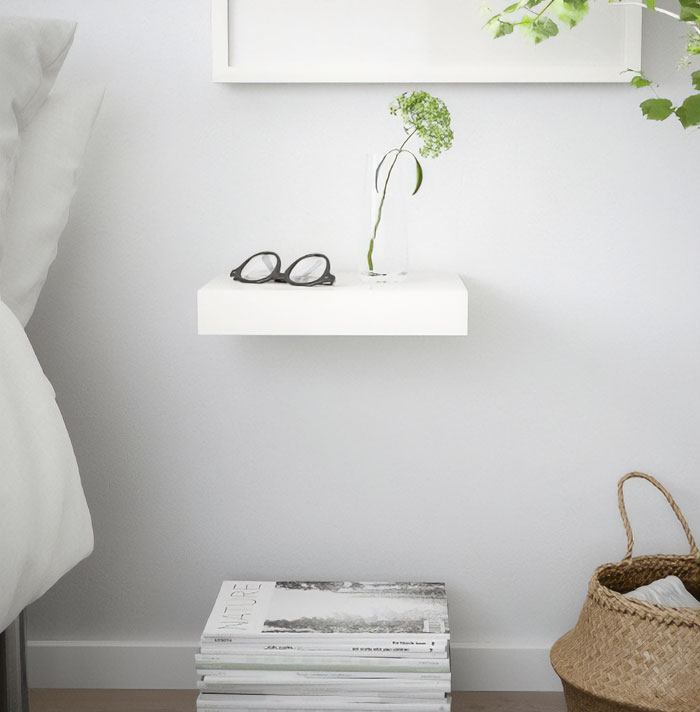 White modern floating nightstand shelf mounted on wall beside bed holding glasses and a small vase with flower.
